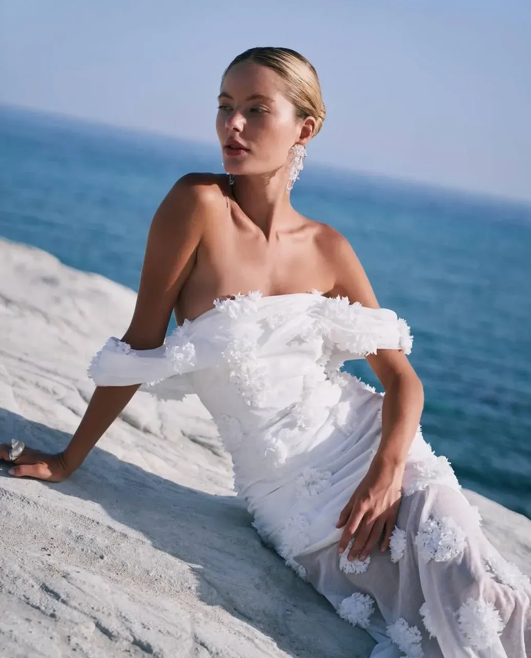 Woman in a white off-shoulder dress standing on a rocky beach by the sea