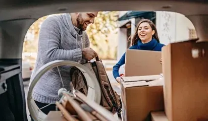 Two people loading a car with boxes and a wrapped item into a car trunk while clearing out belongings