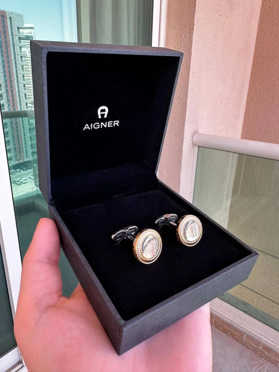 Person holding an AIGNER box with two cufflink-like items, suggesting surplus business gifts that could be given away