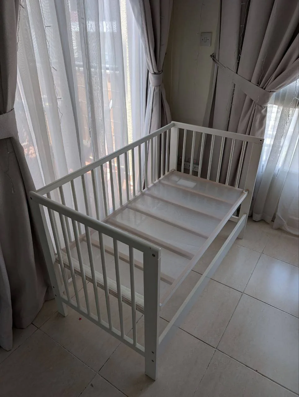 White baby crib in a bright room with sheer curtains and tiled floor