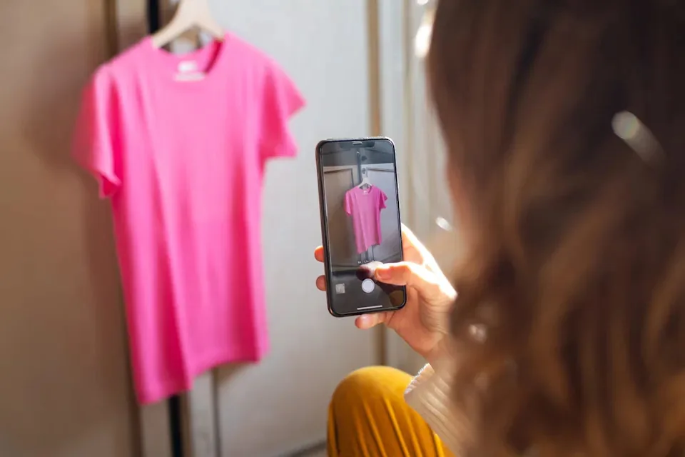 Person photographing a hanging T-shirt with a smartphone to share item details for in-app messaging coordination