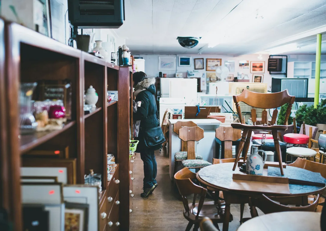 Indoor thrift shop with wooden tables, chairs, and shelves, with a person browsing second-hand furniture