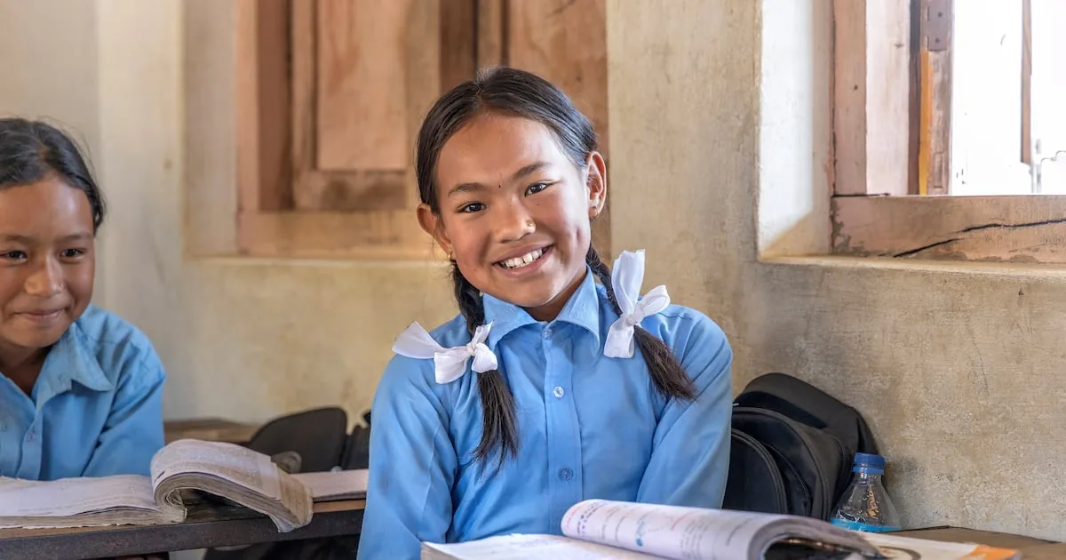 Two schoolgirls in blue uniforms reading and writing at their desks in a classroom