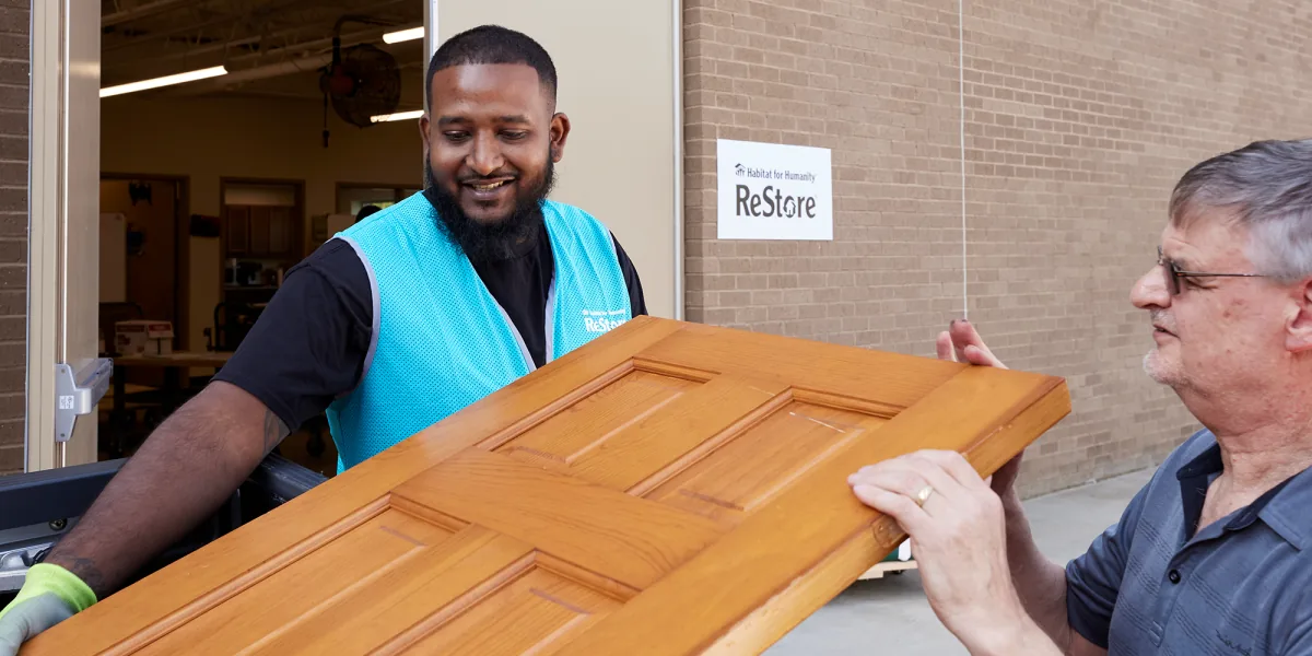 Two men carry a wooden door outside a brick ReStore building
