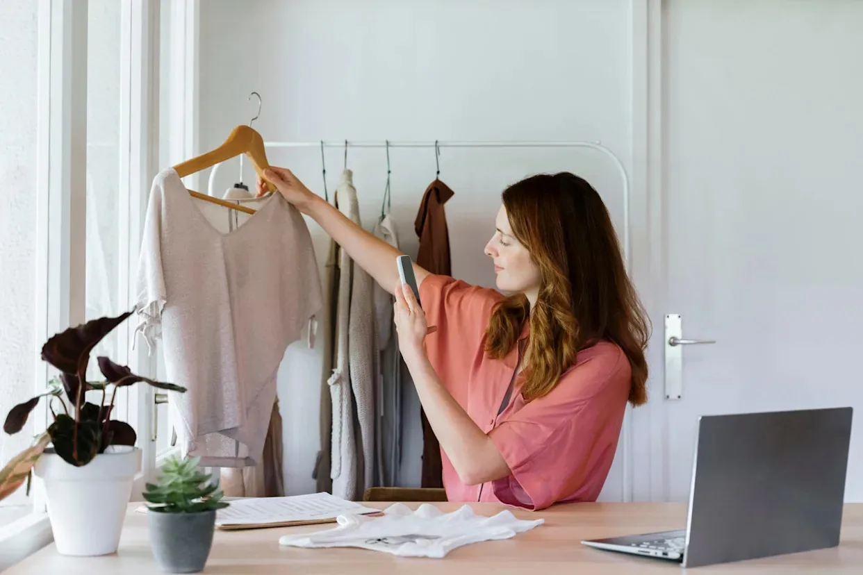 Woman in a small home office area choosing a light sweater from a clothes rack beside her laptop workspace